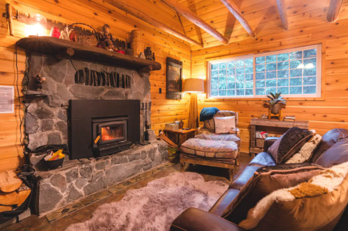 Cozy cabin interior with a stone fireplace, wooden walls, a plush rug, and comfortable seating. Warm and inviting atmosphere.