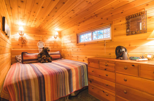 Cozy wooden bedroom with a striped blanket, teddy bears, and a dresser, featuring warm lighting and a small window.