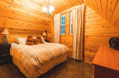 Cozy wooden bedroom with a bed, two teddy bears, lamps, curtains, and a fan, featuring warm, rustic decor.