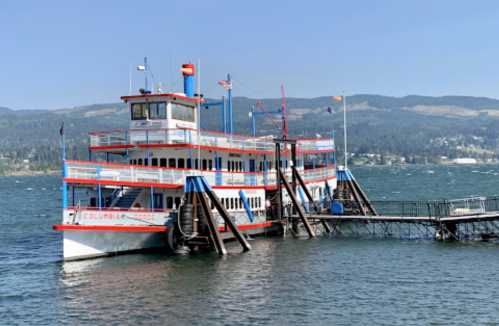 A large riverboat docked at a pier on a calm waterway, surrounded by hills under a clear blue sky.