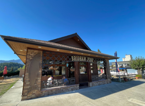A wooden restaurant with a sign reading "Brigham Fish," surrounded by trees and mountains under a clear blue sky.