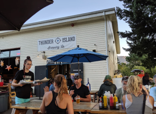 A brewery with outdoor seating, patrons enjoying drinks, and a server bringing food under a blue umbrella.