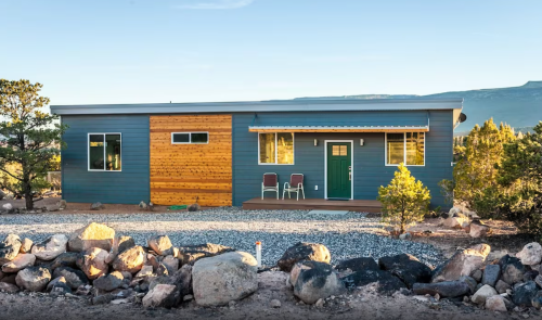 A modern house with a wooden accent wall, surrounded by rocks and greenery, set against a mountainous backdrop.
