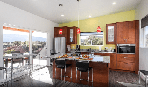 Modern kitchen with wooden cabinets, granite island, red pendant lights, and a view of the outdoors through large windows.