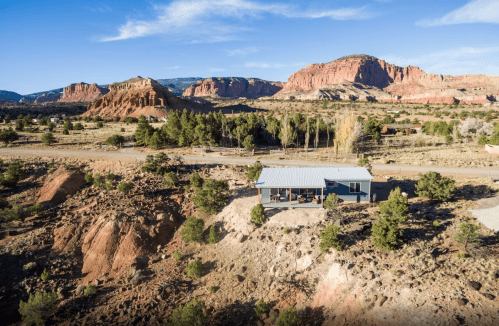 A modern house sits on a rocky landscape, surrounded by trees and mountains under a clear blue sky.