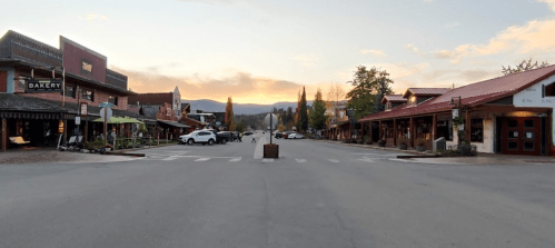 A quiet street in a small town at sunrise, lined with shops and trees, with mountains in the background.