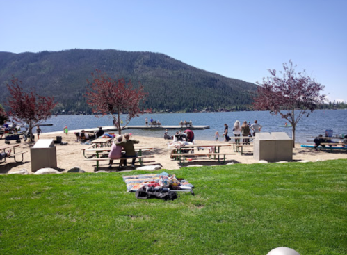 A sunny lakeside scene with people enjoying the beach, picnic tables, and a dock surrounded by mountains.