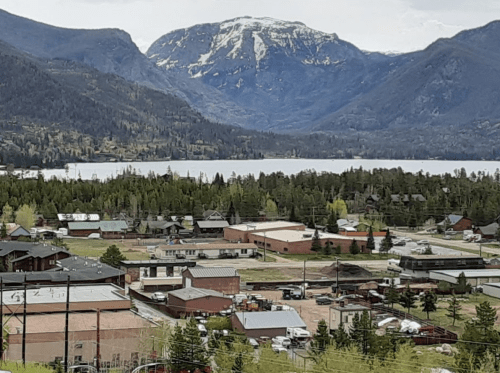 A scenic view of a small town surrounded by mountains and a lake, with greenery and buildings in the foreground.