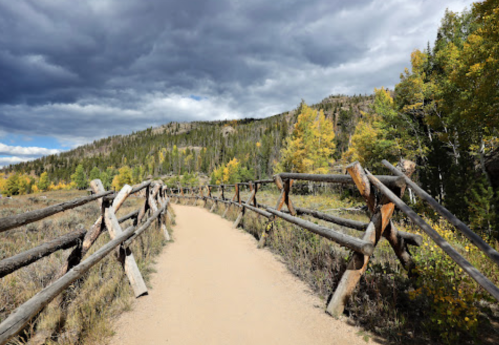 A dirt path bordered by a wooden fence, surrounded by trees and mountains under a cloudy sky.