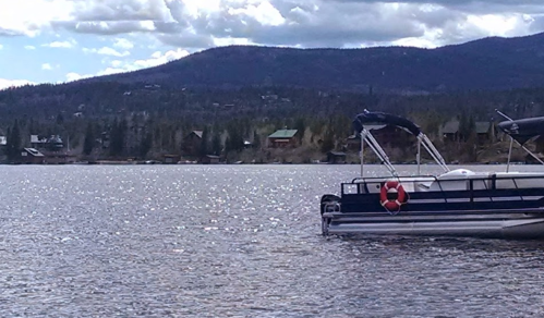 A boat with a life ring is docked on a shimmering lake, surrounded by trees and mountains under a cloudy sky.