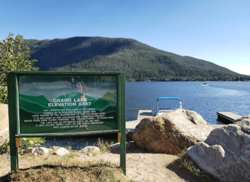 A scenic view of Grand Lake, Colorado, with a sign indicating elevation and a dock by the water surrounded by mountains.
