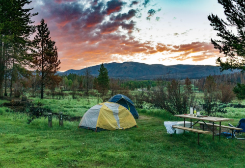 A campsite with two tents, a picnic table, and a scenic view of mountains and colorful clouds at sunset.