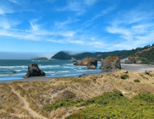 A scenic beach view with rocky formations, gentle waves, and lush green hills under a blue sky with wispy clouds.