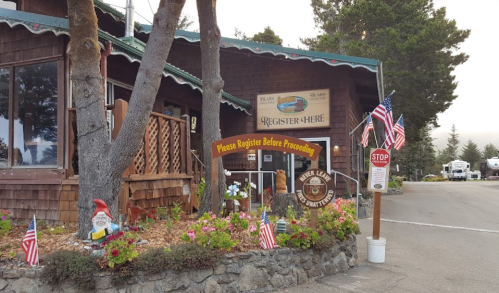 A rustic lodge entrance with a "Register Here" sign, surrounded by flags and a gnome, set in a forested area.