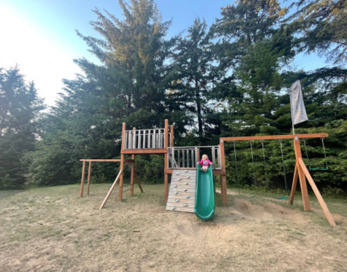 A wooden playground with a slide, swings, and climbing structure, surrounded by trees in a grassy area.
