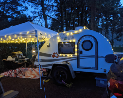A cozy camper trailer with string lights, set up under a canopy in a wooded area during twilight.