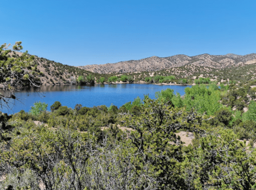 A serene landscape featuring a calm lake surrounded by green trees and rolling hills under a clear blue sky.
