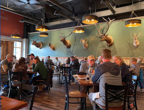A busy restaurant with patrons dining, featuring mounted animal heads on the wall and warm lighting.