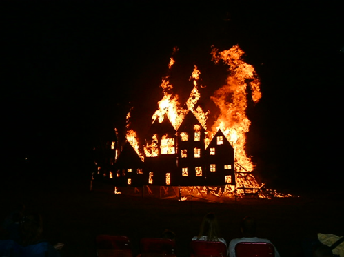 A large wooden structure engulfed in flames, with spectators watching from a distance in the foreground.