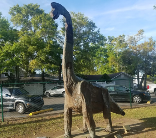 A wooden dinosaur sculpture with a long neck, surrounded by trees and parked cars in the background.