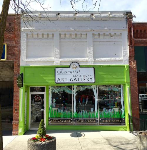 A colorful storefront of "Essential Glass Works Art Gallery" featuring art, chocolate, and fine gifts.