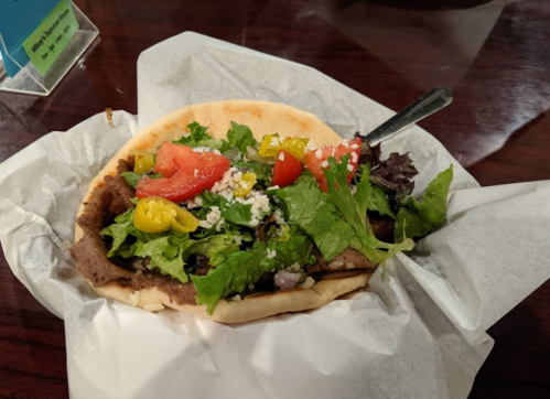 A pita filled with grilled meat, fresh lettuce, tomatoes, peppers, and feta cheese, served on a white paper wrapper.