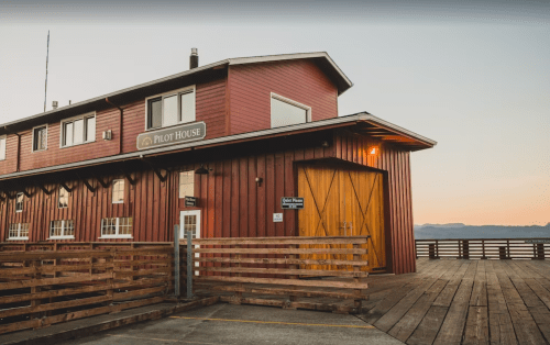 A red wooden building with a sign reading "Pilot House," featuring a deck and sunset in the background.