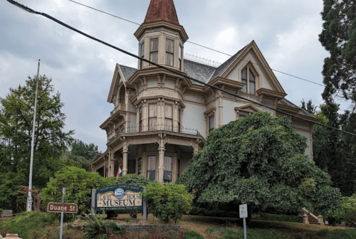 Historic Victorian house with a pointed roof, surrounded by trees, featuring a sign for the "Hatel House Museum."