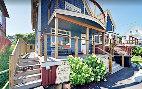 A blue house with a wooden porch, stairs, and signs for private and reserved tenant parking. Lush greenery in front.