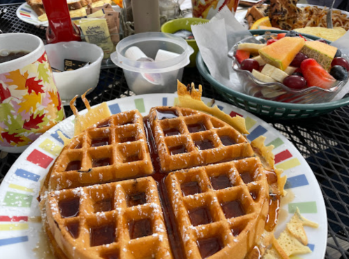 A plate of golden waffles drizzled with syrup, surrounded by a fruit salad and various breakfast items on a table.