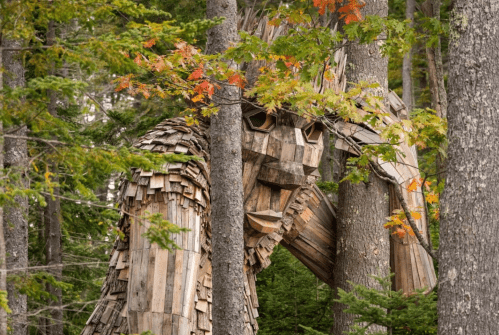 A large wooden sculpture resembling a creature stands among trees, surrounded by autumn foliage.