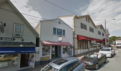 A quaint street scene featuring shops with awnings, parked cars, and a cloudy sky.