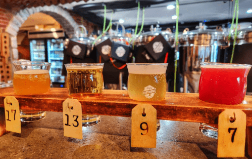 A wooden flight board holds four glasses of different craft beers, labeled with numbers, in a brewery setting.