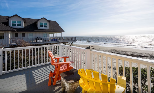 A beachfront view from a deck with colorful chairs overlooking the ocean and a house in the background.