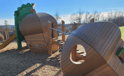 Wooden playground structure with slides and bridges, surrounded by trees and blue sky.