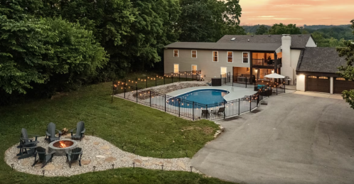 Aerial view of a backyard with a pool, fire pit, and a house surrounded by trees at sunset.