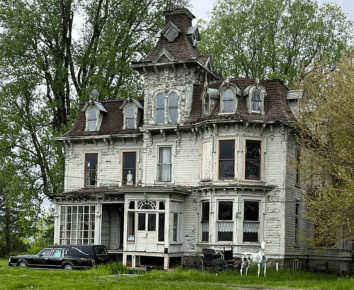 A dilapidated Victorian house with ornate details, surrounded by trees, and an old car parked in front.