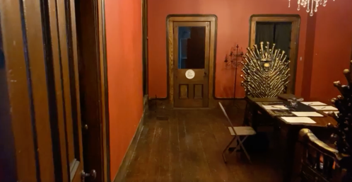A dimly lit room with red walls, featuring a large table, a throne-like chair, and two doors in the background.