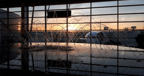A fountain with water jets illuminated by a sunset, set against large windows in an airport terminal.