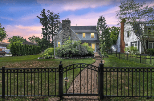 A charming house with a stone chimney, surrounded by greenery and a black iron fence at sunset.