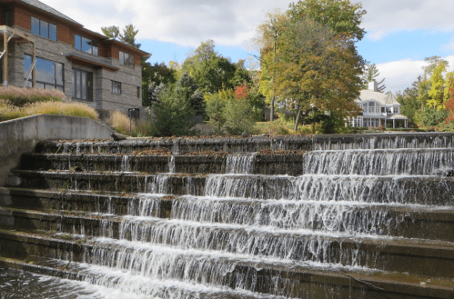 A cascading waterfall flows over stone steps, surrounded by trees and buildings in a scenic outdoor setting.