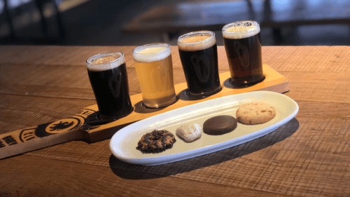 Four glasses of beer in varying colors next to a plate of assorted cookies on a wooden table.