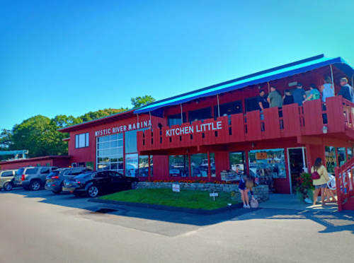 A red building with "Mystic River Marina" and "Kitchen Little" signs, featuring people on a balcony and parked cars.