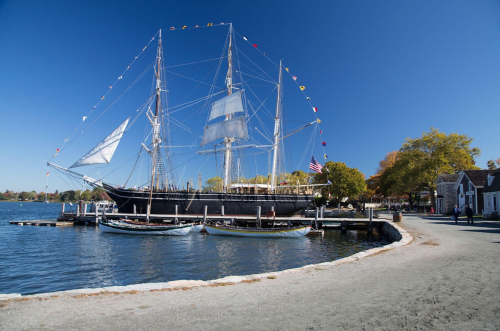 A tall ship with sails raised docked by a sandy shore, with small boats nearby and clear blue skies above.