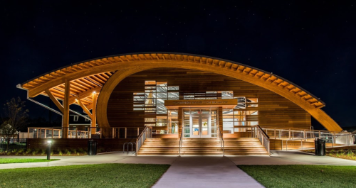 A modern wooden building with a curved roof, illuminated at night, surrounded by grass and a clear starry sky.