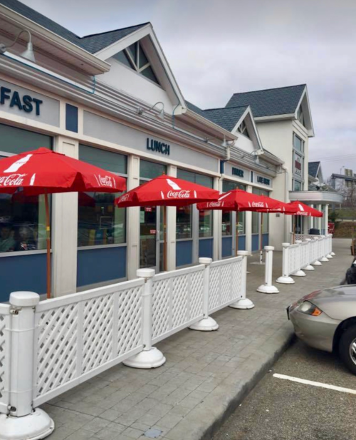 A row of red umbrellas outside a restaurant with white fencing and a parked car nearby.