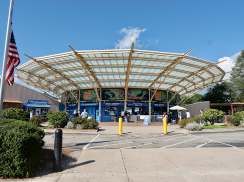 A modern building with a large glass roof, featuring shops and people outside, set against a blue sky.