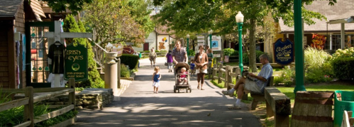 A sunny street scene with families walking, shops lining the path, and greenery in a quaint village setting.