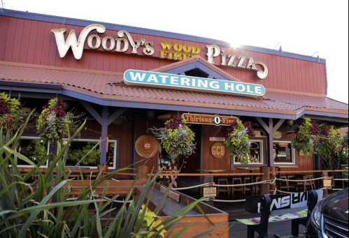 Exterior of Woody's Wood Fire Pizza, featuring a sign for the Watering Hole and vibrant hanging plants.
