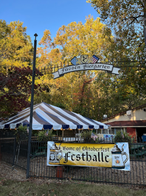 Sign for Füssen Biergarten with a tent and banner promoting Oktoberfest, surrounded by trees in autumn.